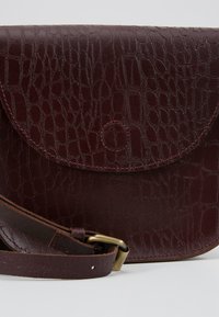 Sac bandoulière en cuir marron foncé avec texture crocodile, rabat incurvé, détail bouton cousu et sangle réglable avec boucle en laiton.