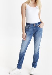 Jeans de denim azul claro con corte ajustado, con cierre de botón, cinco bolsillos y un ligero efecto de desgaste, combinados con una camiseta sin mangas blanca y zapatillas blancas.