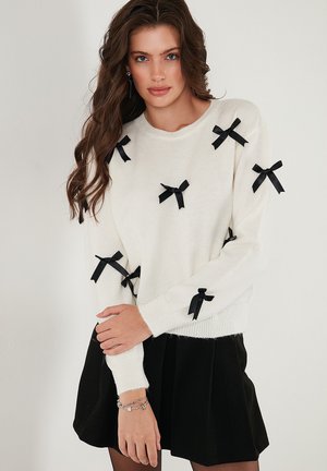 Maglione bianco lavorato a maglia con piccoli fiocchi neri sparsi, scollo rotondo e maniche lunghe, abbinato a una gonna nera plissè.