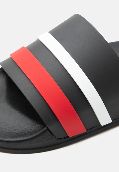 Μαύρο σανδάλι με υφασμένο πέλμα και δύο φαρδιές χρωματιστές λουρίδες, μία κόκκινη και μία λευκή, στο επάνω μέρος.