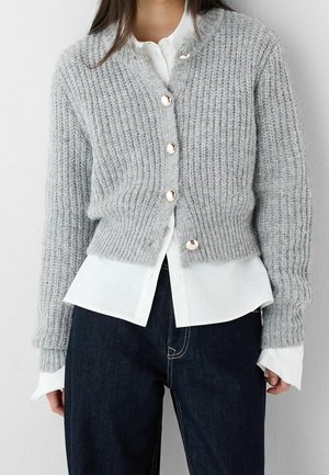 Frau trägt eine hellgraue grob gestrickte Strickjacke mit Perlenknöpfen über einem weißen Hemd und dunklen Jeans.