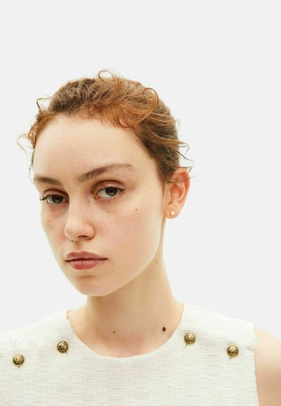 Giovane donna con pelle chiara, lentiggini e capelli rossi mossi raccolti, che indossa piccoli orecchini d'oro e un top senza maniche bianco con trama e bottoni dorati.