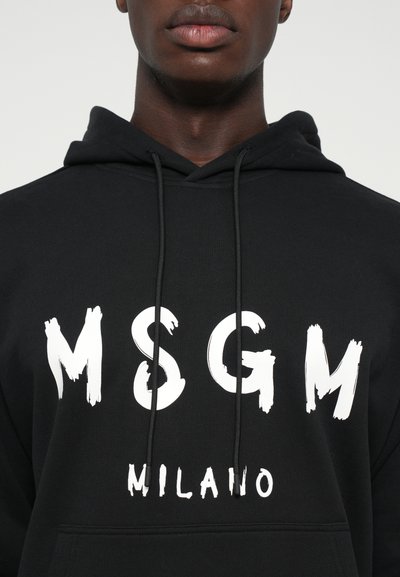 Giovane indossante un felpa nera con il logo vintage bianco "MSGM MILANO" e i lacci neri visibili sul davanti.