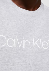 Šedé bavlněné tričko s kulatým výstřihem a výrazným bílým logem "Calvin Klein" na přední straně. Měkká, hladká textura s pohodlným střihem.