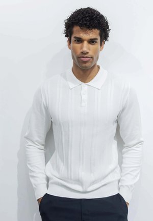 Homme aux cheveux bouclés portant un polo blanc à manches longues et un pantalon foncé, debout devant un fond blanc uni.