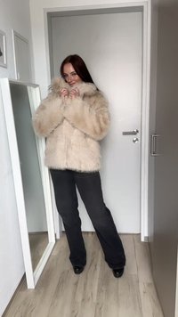 Casaco de pelos sintéticos em bege, com gola alta e acabamento texturizado, combinado com calças pretas e botas de salto, em fundo claro.