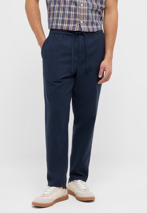 Mann trägt marineblaue Hose mit Kordelzug, beige Sneaker und kurzärmliges kariertes Hemd, steht mit einer Hand in der Tasche vor einfarbigem Hintergrund.