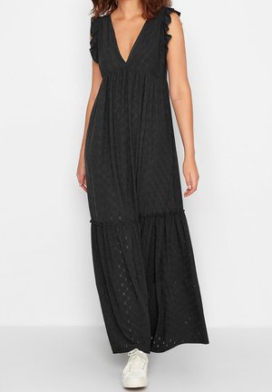 Mujer que lleva un vestido largo negro sin mangas con detalles de ojales y hombros con volantes, combinado con zapatillas blancas, de pie frente a un fondo liso.