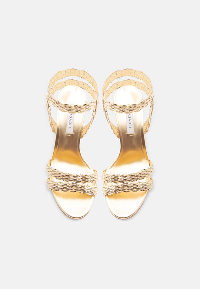 Χρυσά μεταλλικά σανδάλια με πλεγμένους ιμάντες πάνω από τα δάχτυλα και τους αστραγάλους, με επίπεδη σόλα, φαίνονται από πάνω σε λευκό φόντο.
