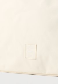 Ύφασμα σε εκρού χρώμα με μικρό ορθογώνιο έμβλημα λογότυπου Lacoste Paris που διαθέτει ανάγλυφο έμβλημα κροκόδειλου στο κέντρο.