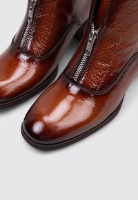 Botas de cuero marrón hasta el tobillo con un acabado brillante, que cuentan con un cierre lateral y una punta redondeada. El tacón apilado añade altura y estabilidad.