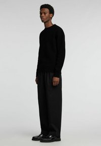 Schwarzer Wollpullover mit Rundhalsausschnitt, kombiniert mit locker sitzenden schwarzen Hosen und glänzenden schwarzen Lederschuhen, stehend vor einem schlichten Hintergrund.