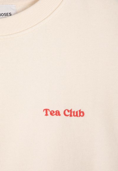 Tissu couleur crème avec encolure côtelée et texte rouge brodé "Tea Club" centré sous le col.