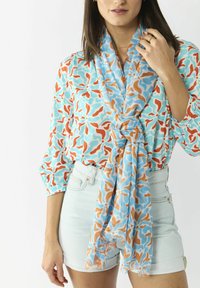 Lichtgewicht sjaal in blauwe, oranje en turquoise patronen, elegant om de nek gedrapeerd. De stof lijkt zacht, met franje randen.