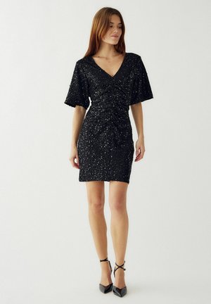Čierne flitrové šaty s výstrihom do V, krátkymi volánovými rukávmi, nariasením vpredu a priliehavým strihom. Vysoké podpätky.