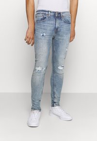 Hombre vestido con jeans skinny desgastados de color azul claro y zapatillas blancas, de pie contra un fondo blanco liso.