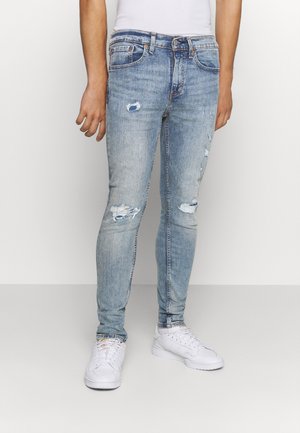 Mann in hellblauen, zerrissenen Slim-Fit-Jeans und weißen Sneakers, der vor einem schlichten weißen Hintergrund steht.