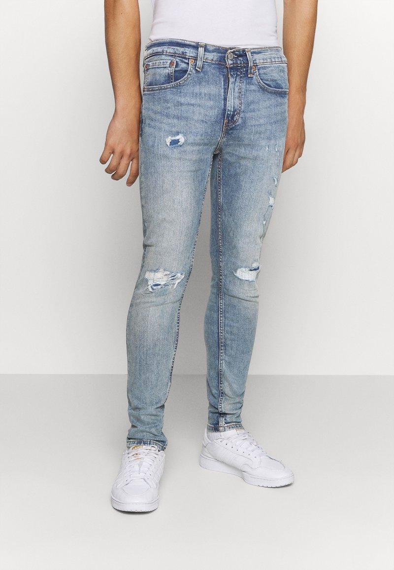 Hombre vestido con jeans skinny desgastados de color azul claro y zapatillas blancas, de pie contra un fondo blanco liso.