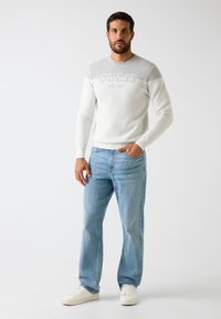 Maglione bianco a maglia con accenti grigi e logo "GUESS EST.1981", abbinato a jeans di denim azzurro chiaro e sneakers bianche.