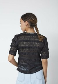 Kvinde med flettet hår, der vender bort, iført en gennemsigtig sort blonde-top med flæse-detaljer og lyseblå denimjeans.