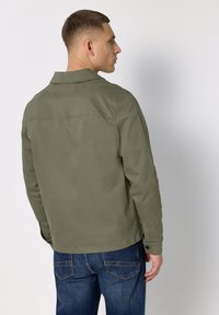 Kurtka w oliwkowej zieleni z kołnierzem, zapinanymi na guziki mankietami i gładką fakturą. Połączona z ciemnoniebieskimi dżinsami z tylnymi kieszeniami.