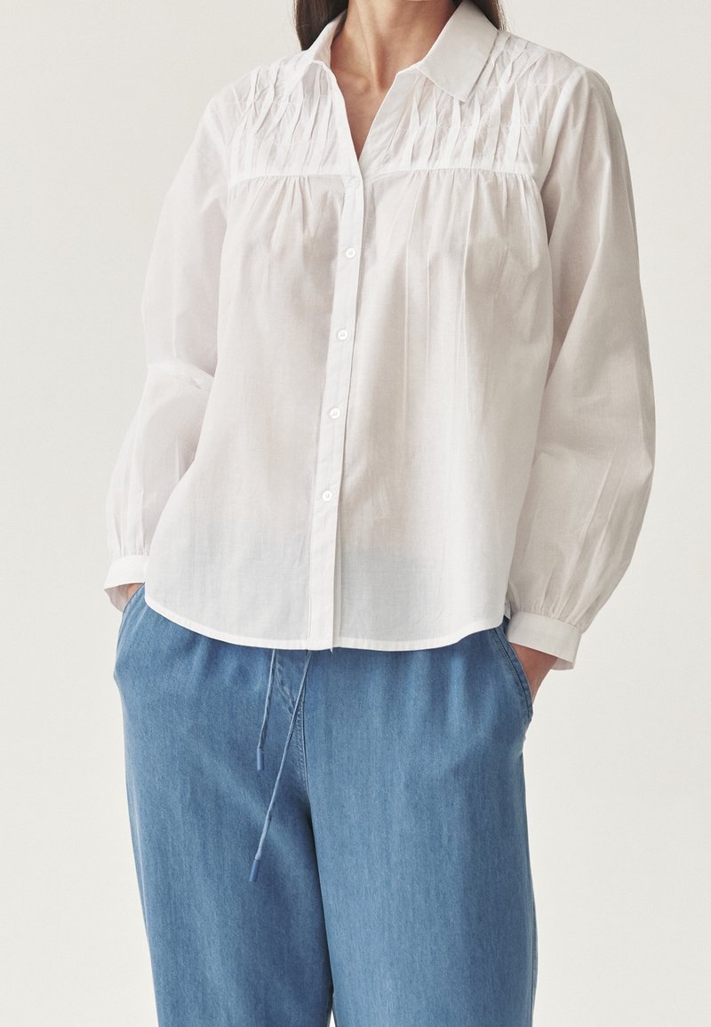 Femme portant un chemisier blanc à manches longues boutonné avec des détails plissés, associé à un pantalon en jean bleu ample. Mains dans les poches.