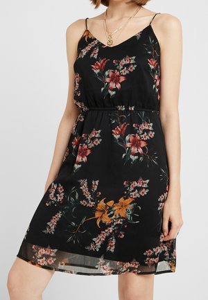 Kobieta w czarnej, kwiatowej sukience na ramiączkach z cienkimi paskami i warstwowymi złotymi naszyjnikami na jasnym, jednolitym tle.