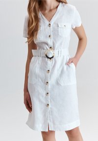 Weißes Leinenkleid mit kurzen Ärmeln, Knopfverschluss vorne, zwei Brusttaschen und einem breiten Gürtel mit einer runden Schnalle. Mittellang mit seitlichen Schlitz.