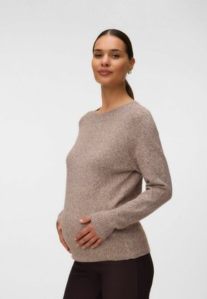 Rase juustega rase naine, kellel on hobusesaba, beige kampsun ja tumedad püksid, õrnalt hoides oma kõhtu, seisab ühevärvilise tausta ees.