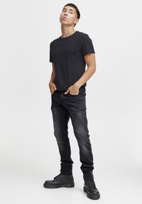 Zwarte t-shirt met korte mouwen, gecombineerd met donkerdenim jeans en zwarte veterschoenen, staand met de handen in de zakken tegen een effen achtergrond.