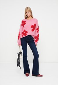 Suéter rosa tricotado com padrões florais vermelhos, combinado com jeans flare azul escuro. Os acessórios incluem um lenço de bolinhas e uma bolsa branca.