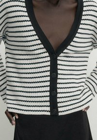 Cardigan rayé noir et blanc en tissu tricoté, avec un décolleté en V profond, fermeture à boutons noirs et manches longues.