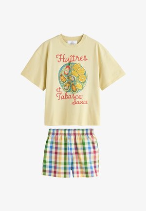 Beiges kurzärmliges T-Shirt mit einer Illustration von Austern, Zitrone und Tabasco-Sauce sowie rotem Text, kombiniert mit mehrfarbigen karierten Shorts.