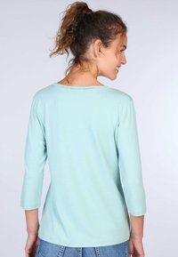 Hellblaues Oberteil mit drei Viertelärmeln, weitem Halsausschnitt und dezenten Nähten entlang des Kragens. Weicher Stoff mit lockerer Passform.