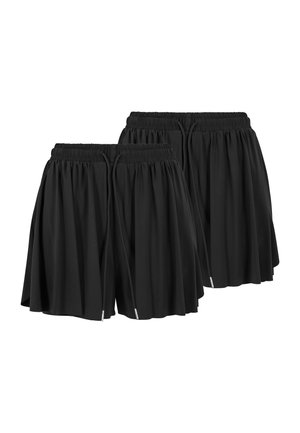 Femme aux cheveux bouclés portant un t-shirt blanc court ajusté et un short noir plissé large, debout devant un fond gris uni.