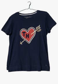 Mörkblå T-shirt med korta ärmar, med en grafik av ett röd hjärta och pilar med guldaccenter och bokstäverna "TH" i mitten.