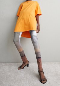 Oranžové oversized tričko, šedé a hnědé vzorované legíny a černé sandály na vysokém podpatku. Materiál trička vypadá jako bavlna, legíny jsou syntetické.
