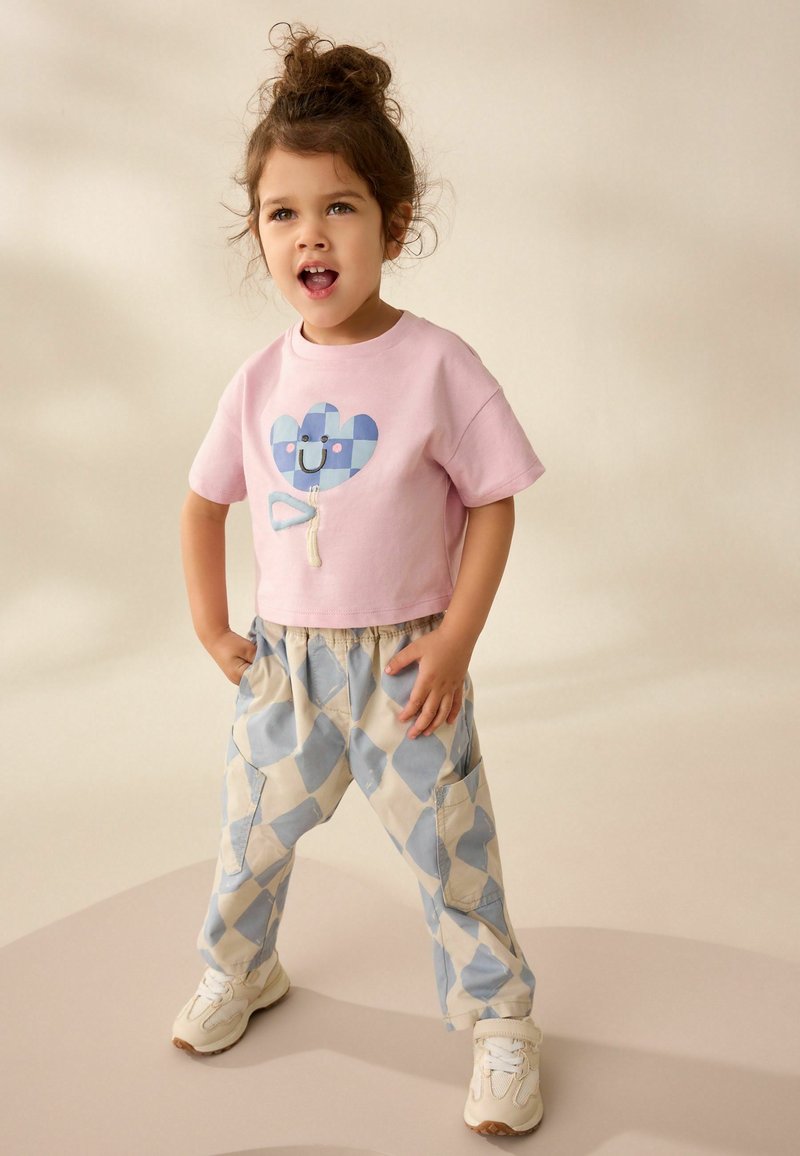 Bambino piccolo con capelli ricci, maglietta rosa e pantaloni blu a quadri, in piedi con le mani in tasca, bocca aperta, su uno sfondo uniforme.