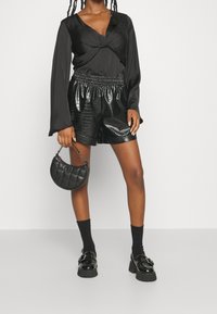 Haut en satin noir à manches longues avec un devant noué, associé à un short noir brillant à motif crocodile, des chaussettes noires aux chevilles et des chaussures noires épaisses.