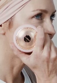 Dispositif facial en silicone tenu contre la joue, présentant une surface texturée avec un motif doré et un boîtier extérieur rose avec un logo noir.
