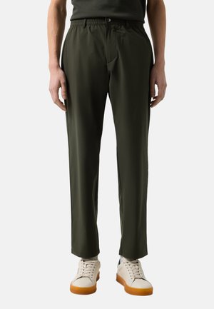 Mann trägt dunkelolivgrüne, gerade geschnittene Hose mit elastischem Bund und weiße Sneaker mit Gummisohlen, steht vor einfarbigem Hintergrund.