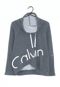 Šedá mikina s dlouhými rukávy a bílým nápisem "Calvin". Má vysoký límec, šňůrky a šikmý lem z měkké, texturované látky.