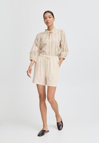 Blusa a righe beige con maniche a palloncino e scollatura a chiave, abbinata a pantaloni beige chiaro e scarpe slip-on scure.