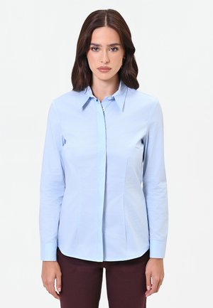 Femme aux cheveux bruns portant une chemise bleu clair à manches longues boutonnée et un pantalon foncé, debout sur un fond blanc uni.