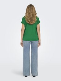 Hosszú hullámos szőke hajú nő zöld rövid ujjú ingben és világoskék bő szárú farmernadrágban, háttal áll egy egyszínű háttér előtt.