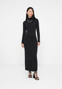 Czarna, długa sukienka maxi z długim rękawem, dopasowana i z wysokim dekoltem, wykonana z elastycznego materiału. Modelka nosi czarne sandały na obcasie z paskami oraz naszyjnik z pereł.