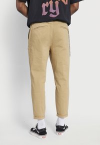 Khaki chinos slim fit, lahkeet həmm, tərəf cibləri və metal zəncir detalları ilə. Ağ corablar və qara idman ayaqqabıları ilə geyinilib.
