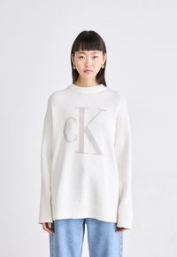 Bela pletena pulover s poudarjenim bež "CK" logotipom na sprednji strani, oversize kroj, ribana ovratnik in široki rokavi, v kombinaciji z svetlo modrimi kavbojkami.