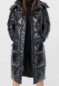 Černá lesklá péřová bunda s kapucí, se zapínáním na zip a prošívaným designem. Má boční kapsy a mírně oversize střih.