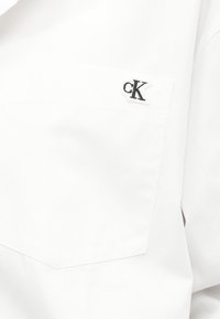 Λευκό ένδυμα με μπροστινή τσέπη που έχει ραμμένο ένα μικρό ασπρόμαυρο λογότυπο "cK" κοντά στο πάνω άκρο της τσέπης.